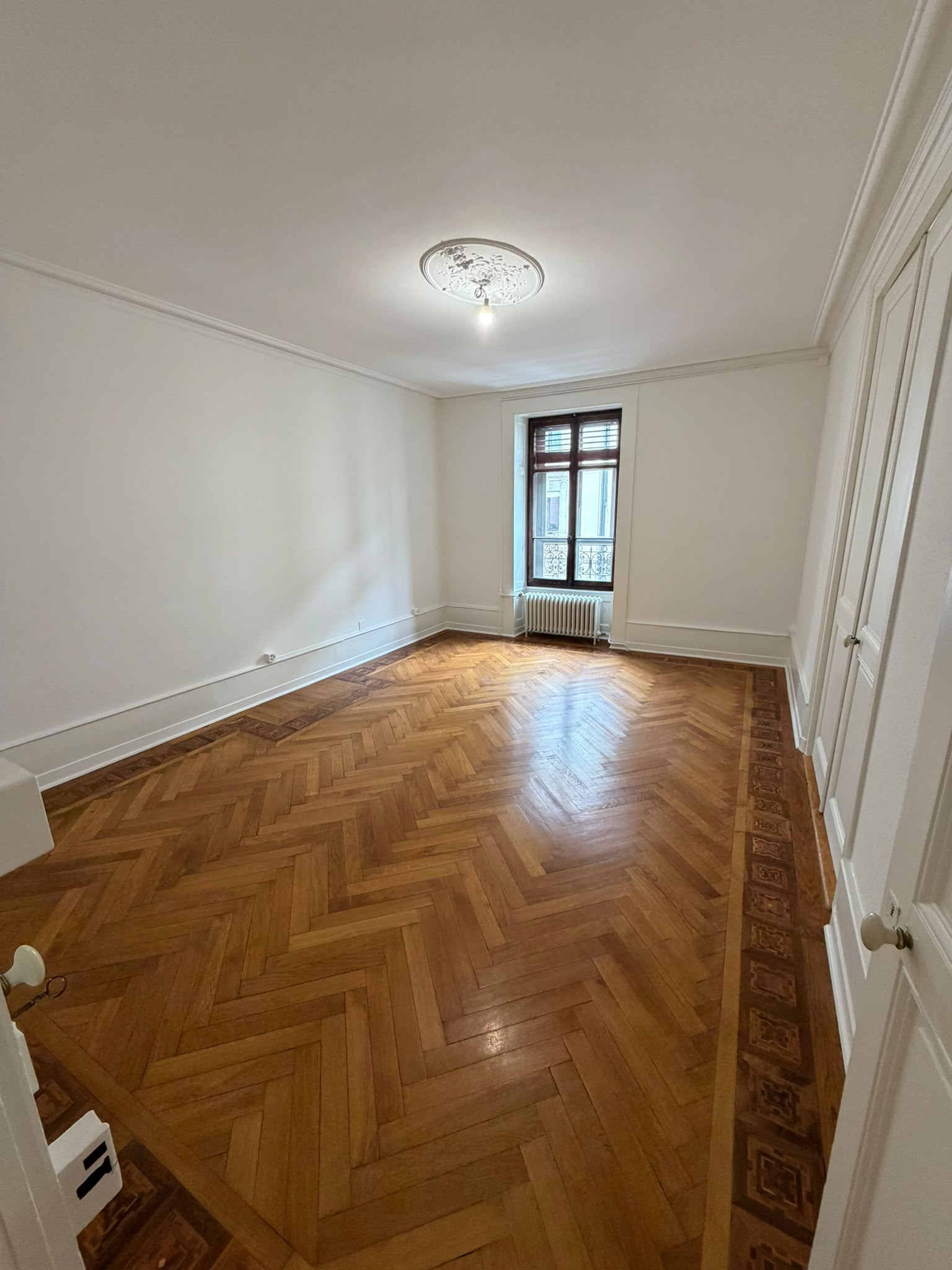 Chambre repeinte en blanc dans un appartement ancien à Genève : plafond avec rosace, murs lisses, parquet à bâtons rompus